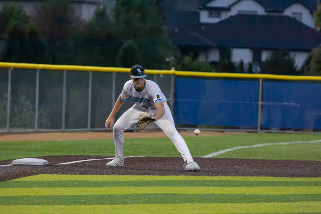 A baseball player in a white uniform with a cap crouches near first base, preparing to field a ground ball on the infield during a game in a dimly lit park.