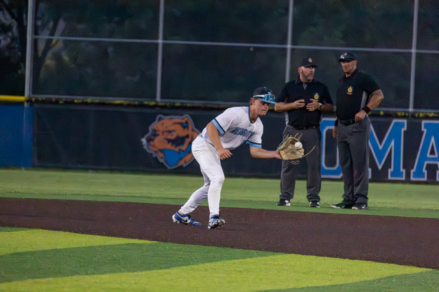A baseball player in a white uniform prepares to field a ball on an infield dirt area, with umpires visible in the background.