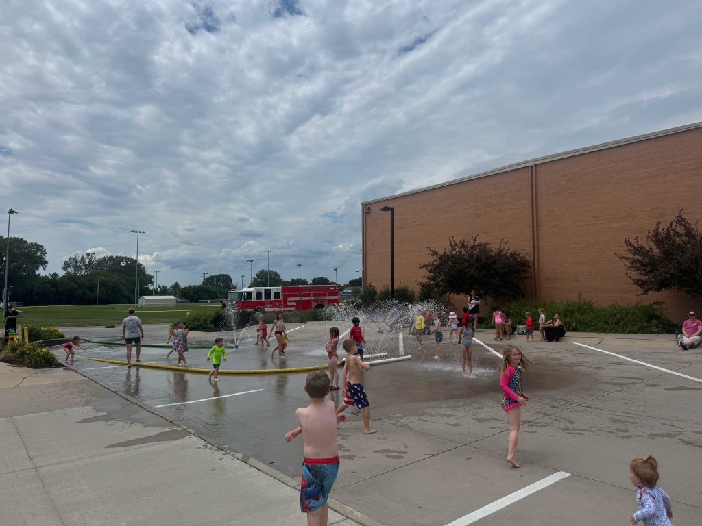 Children playing in a water spray area on a sunny day, with a fire truck visible in the background.