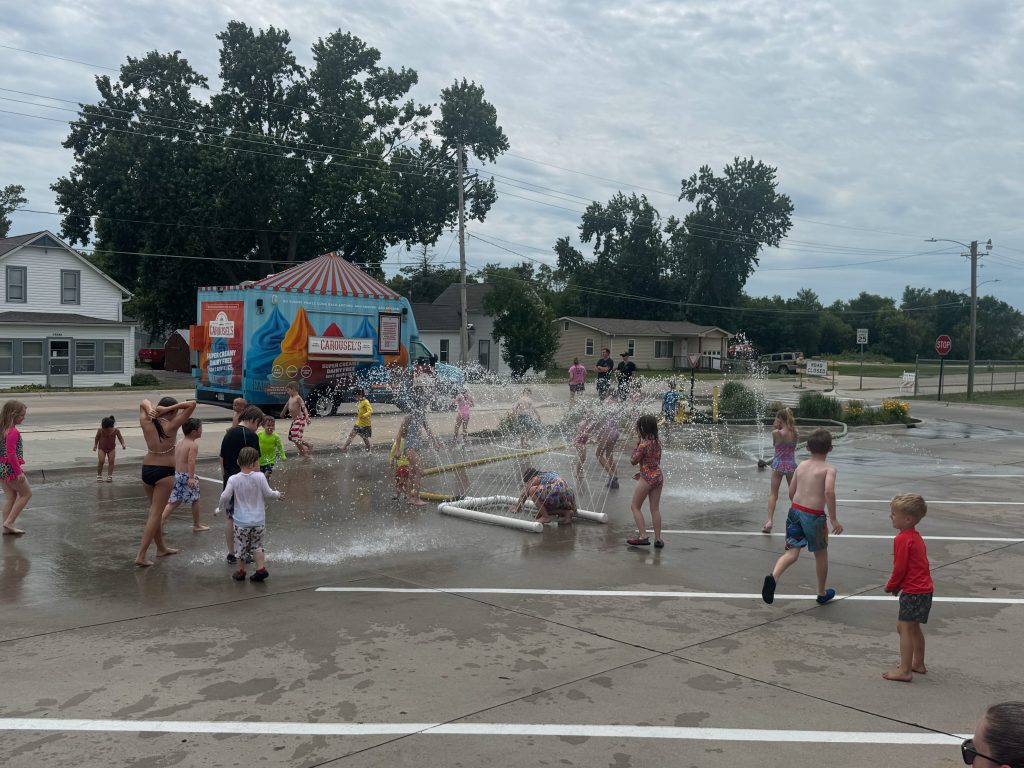 A group of children playing and splashing in water outside near a colorful ice cream truck, with some kids in swimsuits enjoying the summer day.