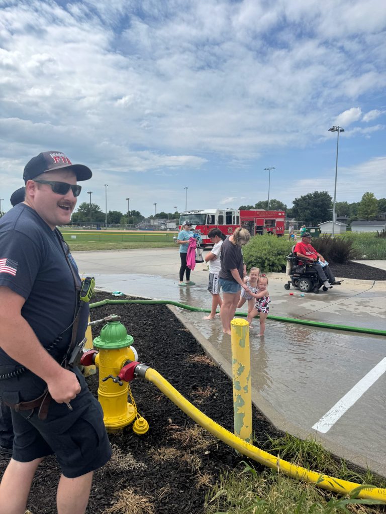 A firefighter smiles while supervising children playing in water from a fire hydrant, with a fire truck in the background and spectators nearby.