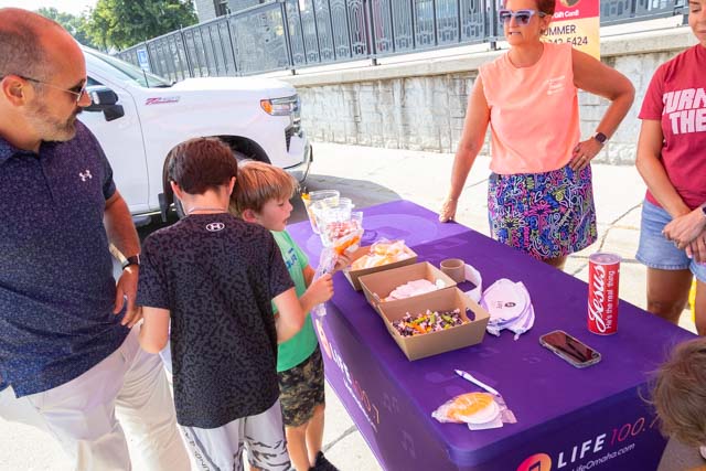 A group of people, including two children, interacting at a table with snacks and drinks outdoors. One adult woman is serving food while others observe, with a truck parked in the background.