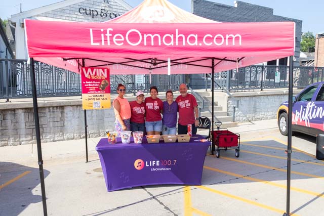 Group of five people standing behind a table under a red canopy with the website LifeOmaha.com displayed, promoting a local event with snacks and beverages.