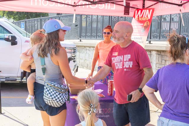 A woman with a child on her back shakes hands with a man at a vendor booth under a tent, while another woman in the background watches.