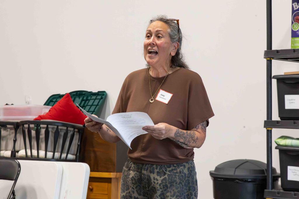 A woman with long hair and tattoos enthusiastically speaks while holding a paper, wearing a brown top and patterned pants in a well-lit indoor setting.