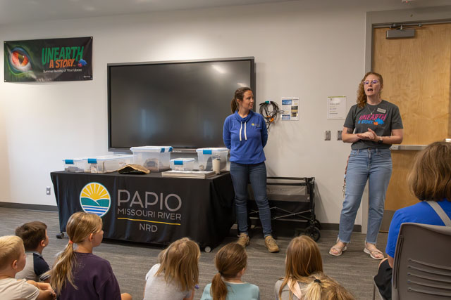 Two women speaking in front of a group of children in a classroom setting, with a display promoting the Papio Missouri River NRD and the theme 'Unearth a Story.'