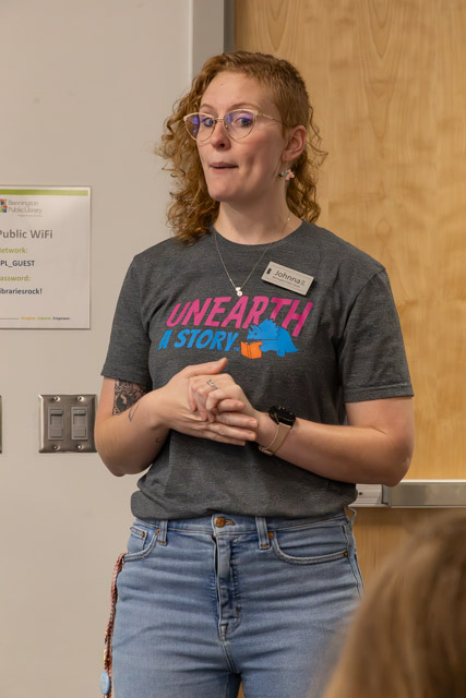 A person with curly hair and glasses stands in front of a wooden door, wearing a gray t-shirt that reads 'UNEARTH A STORY.' They appear to be speaking or presenting.