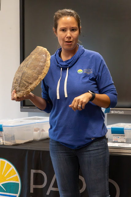 A woman in a blue hoodie holding a large turtle shell while speaking to an audience.