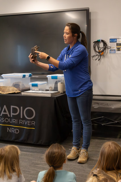 A presenter holds a turtle shell while engaging with an audience of children in a classroom setting.