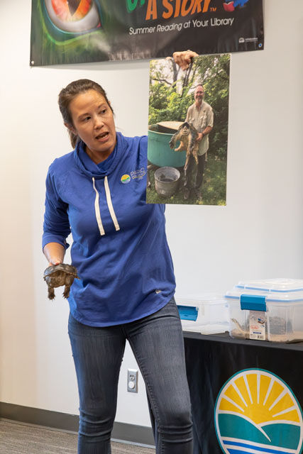 A woman in a blue hoodie holding a turtle in one hand while displaying a photo of a man holding a turtle on a poster during a presentation.