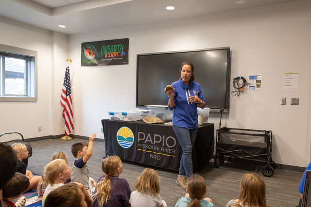 An educator speaks to a group of children seated on the floor during a presentation in a classroom, with a table displaying educational materials and an American flag visible in the background.