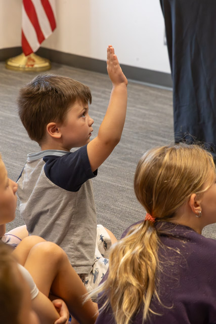 A young boy raising his hand in a classroom setting, sitting among other children on the floor.