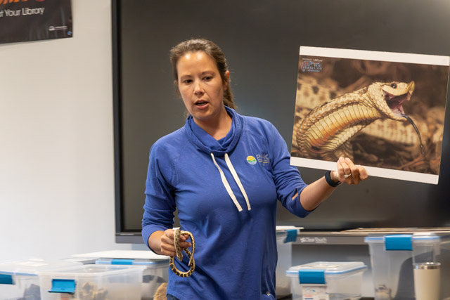 A woman in a blue hoodie holding a snake in one hand and a picture of a snake in the other, speaking to an audience in an educational setting.
