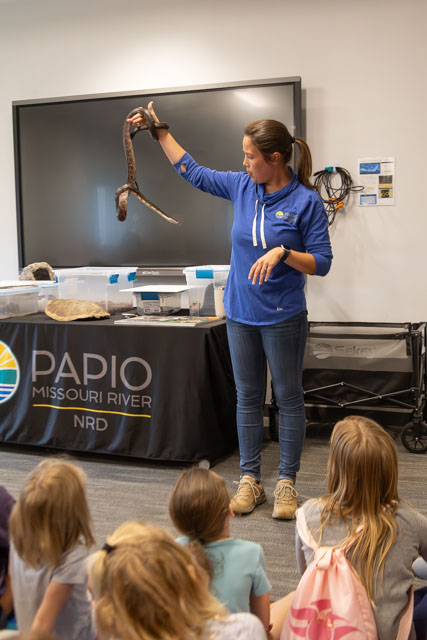 A presenter holding a snake while giving a demonstration to an audience of children seated on the floor, with a display table and a logo in the background.