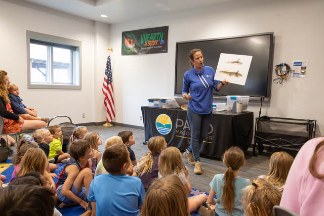 A presenter holds an illustration of marine life while speaking to a group of children seated on the floor in a classroom setting.