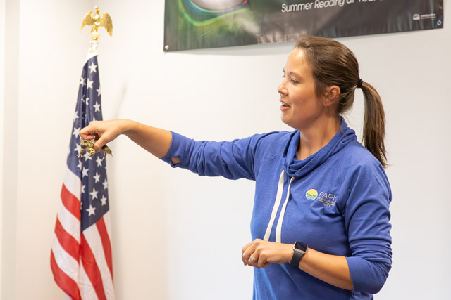 A woman in a blue sweatshirt demonstrates something while holding an object in her hand, with an American flag in the background.