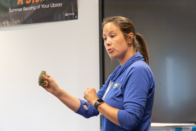 A woman in a blue shirt gestures while holding a small object, possibly an animal, in a library setting.