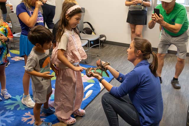 A group of children watching as a woman kneels and holds a snake, demonstrating it to a young girl. Other children are present, showing curiosity and excitement.