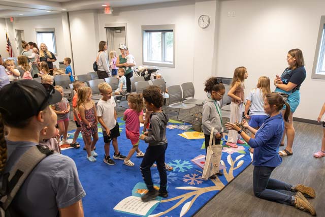 A group of children participating in an activity in a room with chairs and a colorful carpet, while adults supervise and interact with them.
