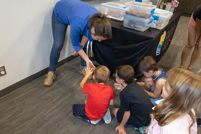 A facilitator in a blue shirt shows an animal to a group of attentive children sitting on the floor, with containers of specimens visible on a table in the background.