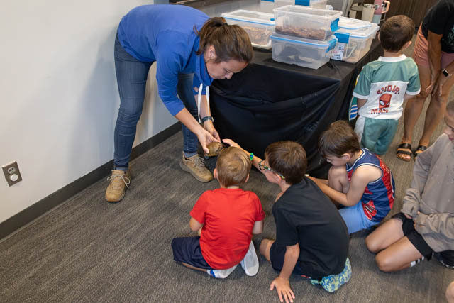 A teacher crouches down to show something to a group of fascinated children sitting on the floor, with storage containers visible in the background.