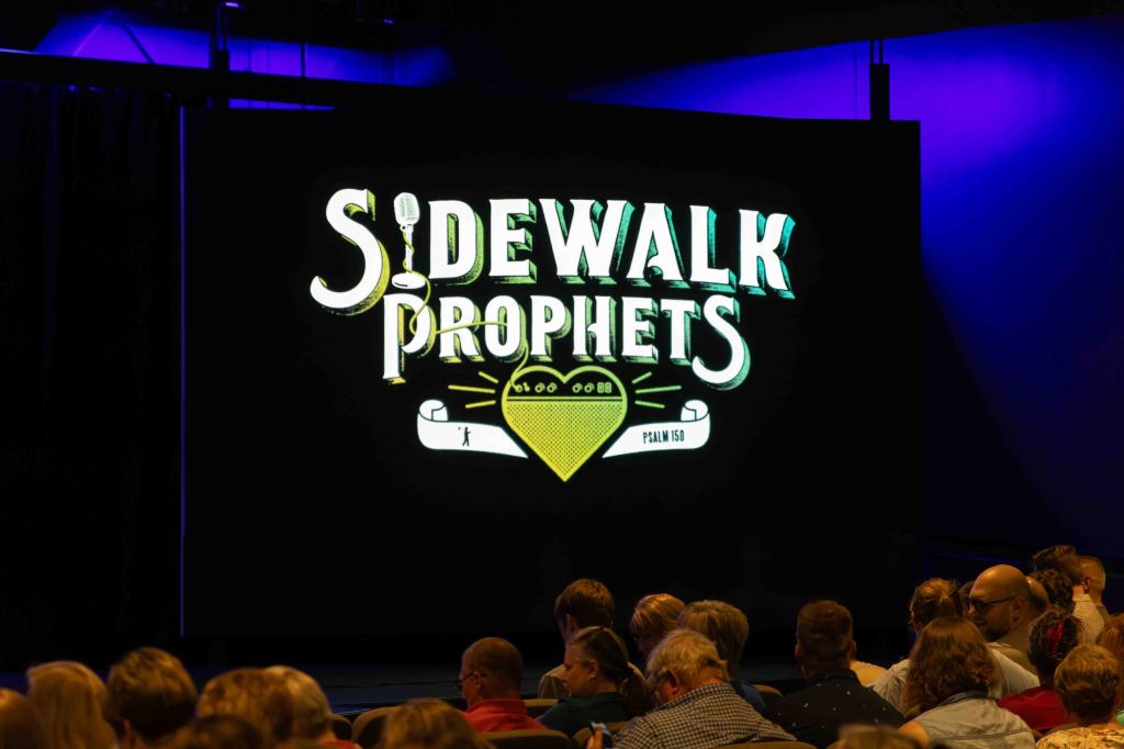Stage backdrop featuring the logo of 'Sidewalk Prophets' with a microphone and a decorative design, in front of an audience.