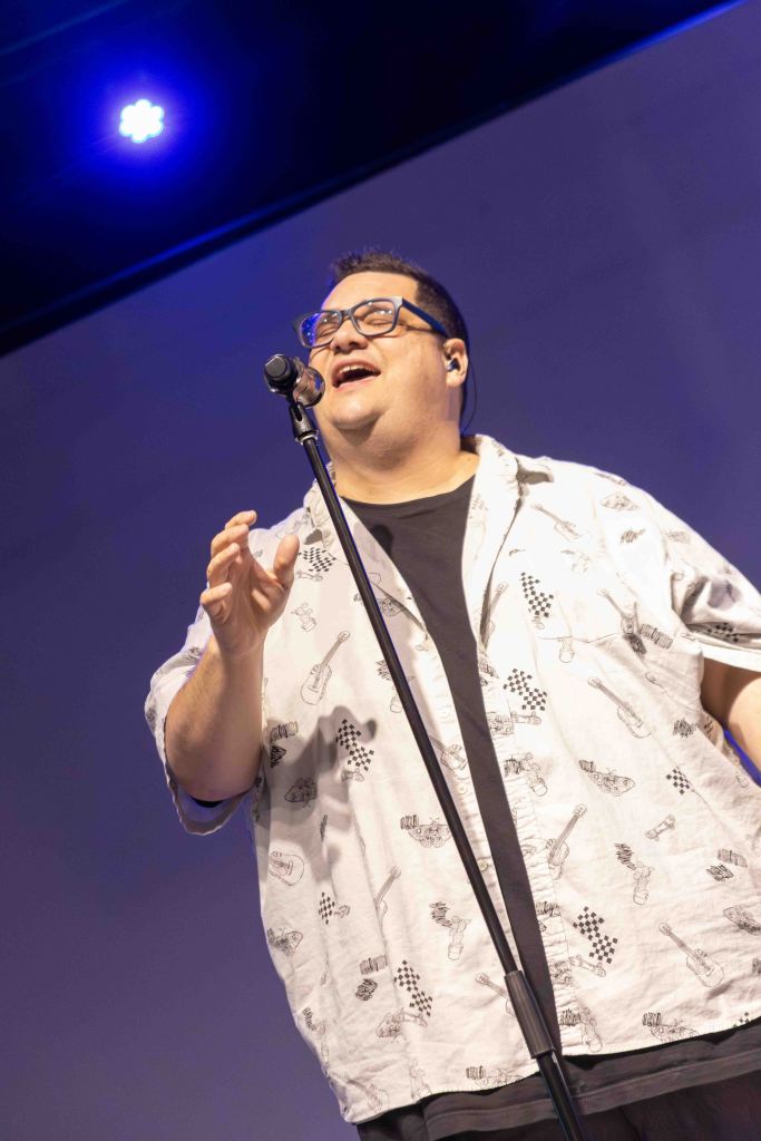 A performer singing on stage with a microphone, wearing glasses and a patterned shirt, engaging the audience.