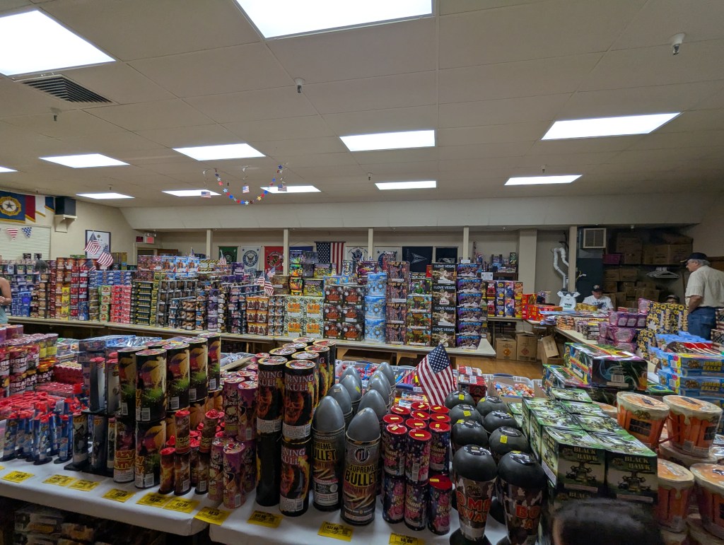 An indoor fireworks store with shelves stocked full of fireworks and decorative items, including small flags and colorful packaging, with customers browsing.