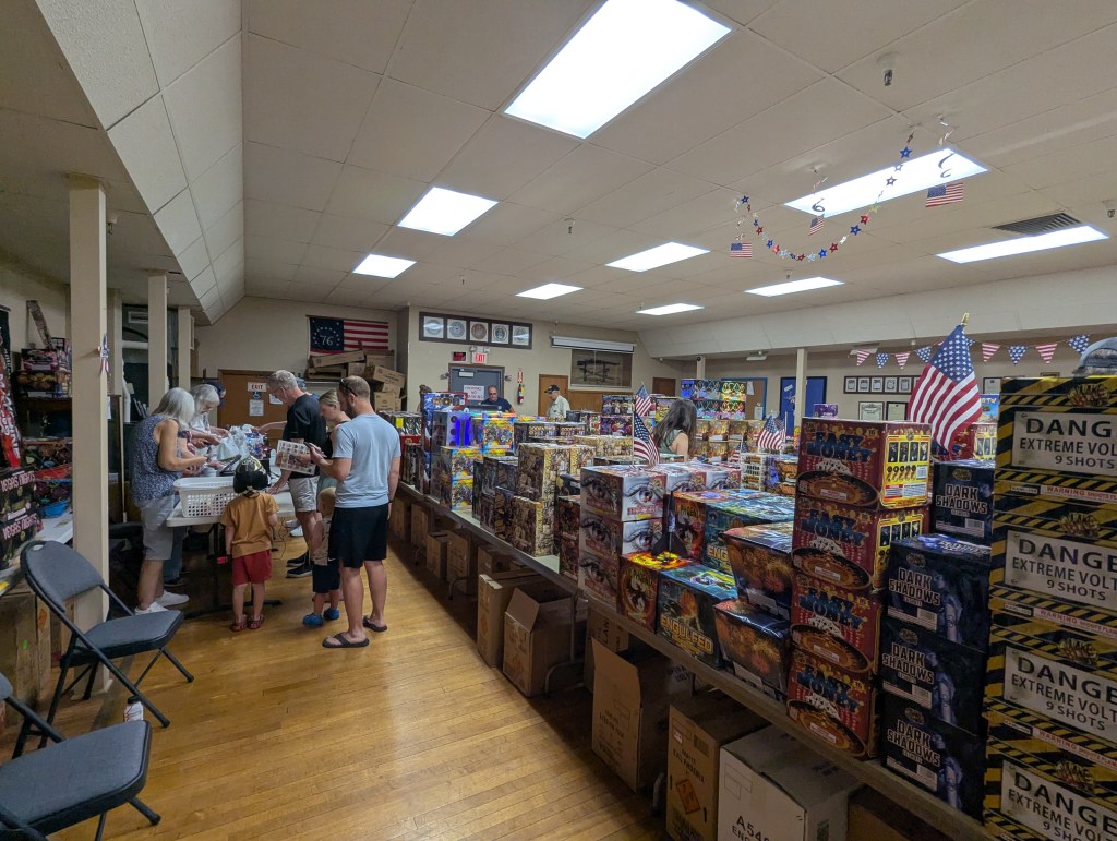 Interior view of a store selling fireworks, with several customers browsing and purchasing items, patriotic decorations visible.