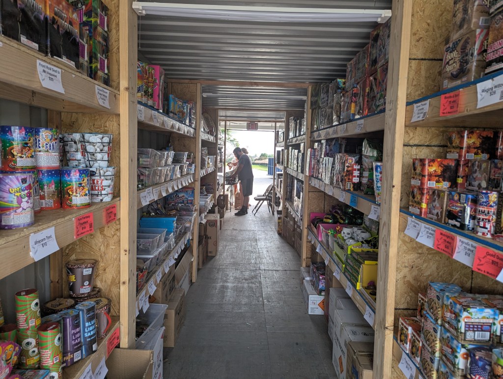 Interior of a fireworks store with packed shelves displaying various fireworks products and a customer browsing.