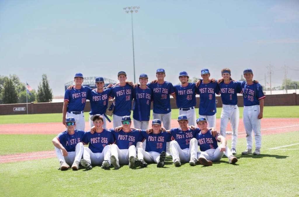 A youth baseball team posing on the field, wearing blue jerseys with 'POST 266' printed on them. The team members are arranged in two rows, smiling and showcasing their uniforms.