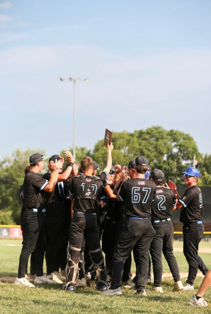 A group of young baseball players in black uniforms celebrating and holding up a trophy on a sunny day.