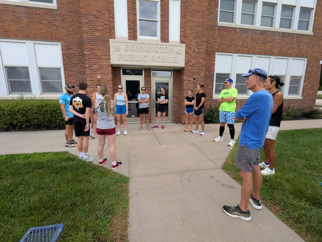 A group of people gathered outside a public school, listening to a speaker in front of the building. The participants are wearing athletic clothing, suggesting a running or fitness event.