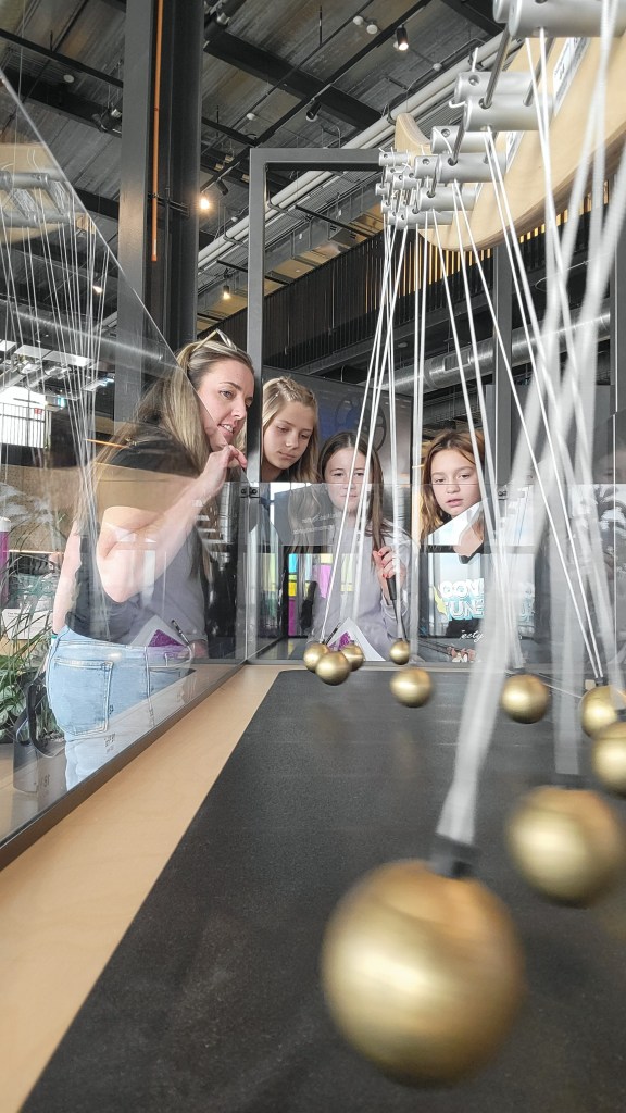 A group of four young girls and a woman are observing a pendulum exhibit in a science museum, with golden pendulums swinging in the foreground.