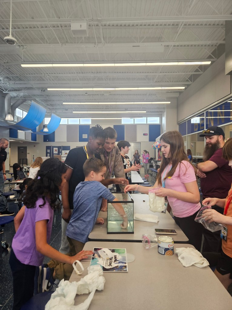 Group of children and adults gathered around a table, observing and participating in an interactive science demonstration involving a fish tank and various materials.