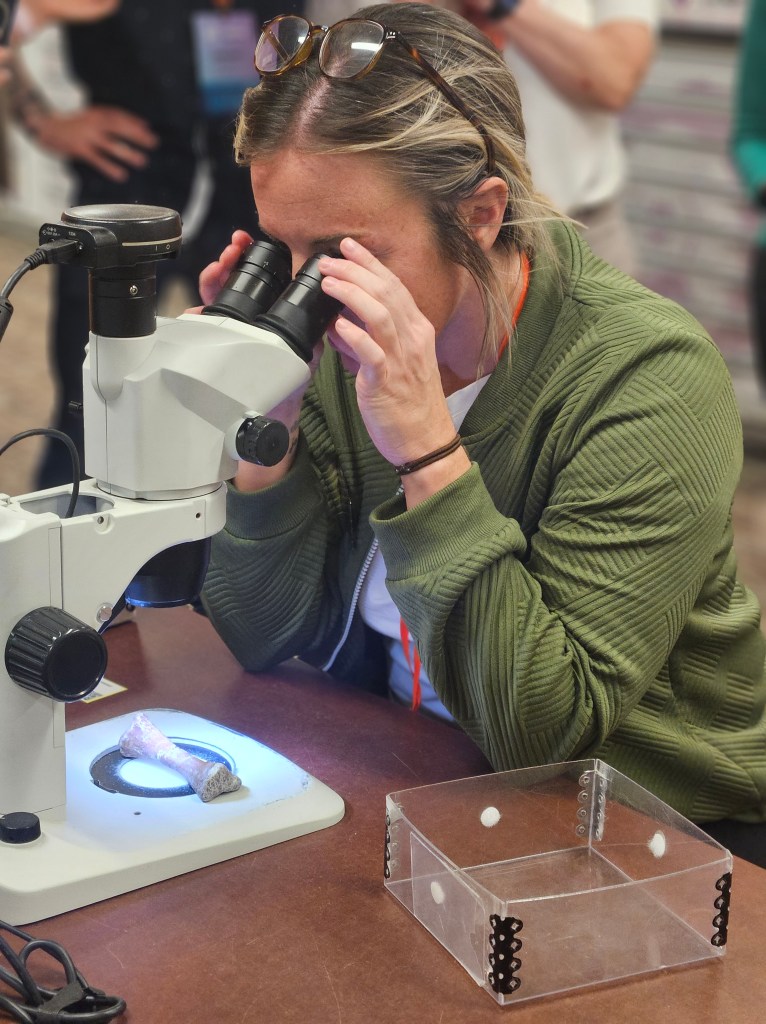 A woman wearing glasses examines a specimen under a microscope, focusing intently. A clear box is visible on the table beside her.