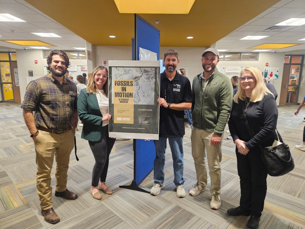 Group of five people standing in a school hallway holding a poster for 'Fossils in Motion: A Student Museum'.