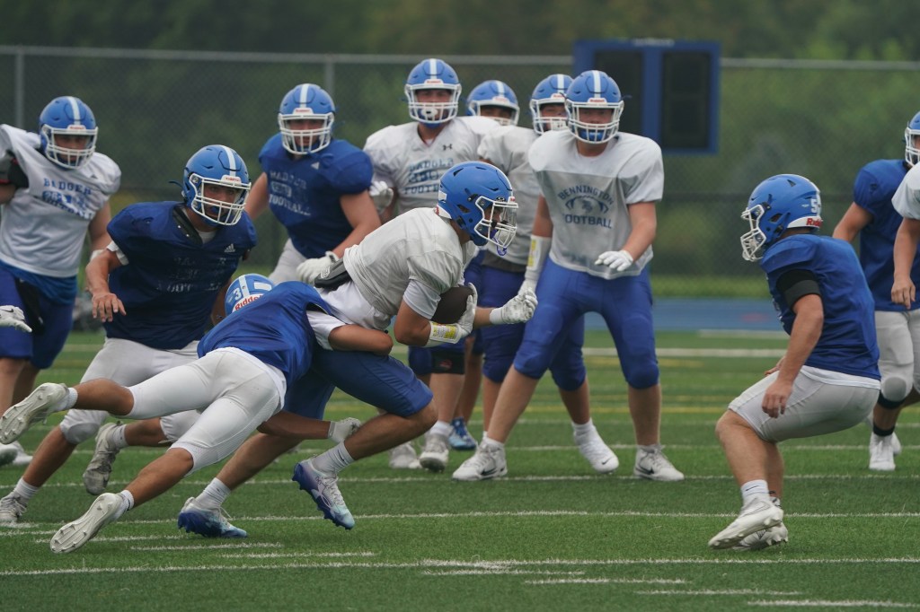 A high school football practice scene with players in blue and white uniforms, one player is being tackled as they run with the ball, surrounded by teammates and opponents on a grass field.