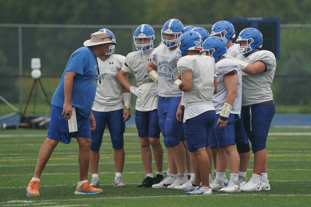 A football coach discusses strategy with a group of players during practice on a grassy field.