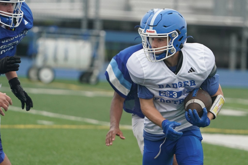 A young football player in a blue and white jersey runs with a football, evading a defender during practice on a grassy field.