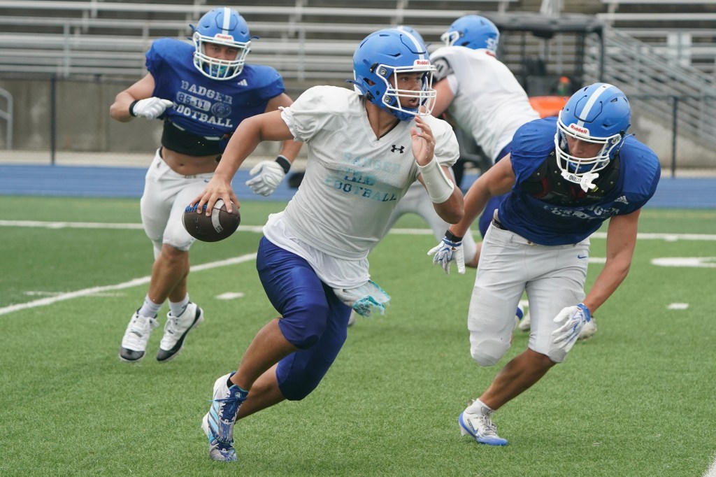 A high school football player in a white jersey scrambles with a football in hand as two defenders in blue jerseys pursue him during practice on a grass field.