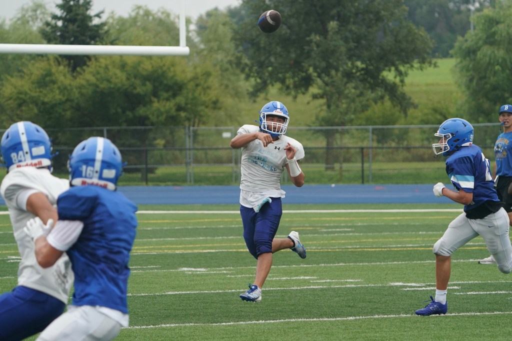 A youth football player in a blue helmet throws a football while other players in blue jerseys are positioned nearby on a grassy field.