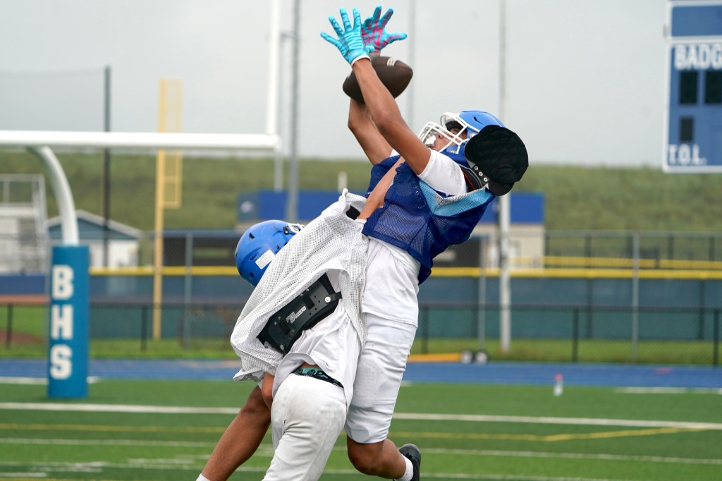 A football player makes an athletic catch while another player attempts a tackle during practice on a field.