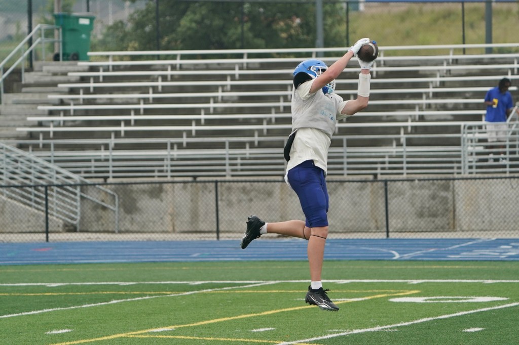 A football player wearing a helmet and jersey jumps to catch a football during practice on a sports field.