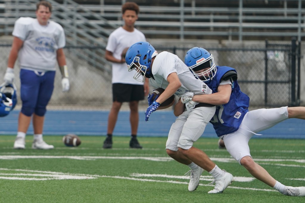 A football player in a blue helmet tackles a player in a white jersey, during a practice session on a grassy field. Other players in the background are watching.