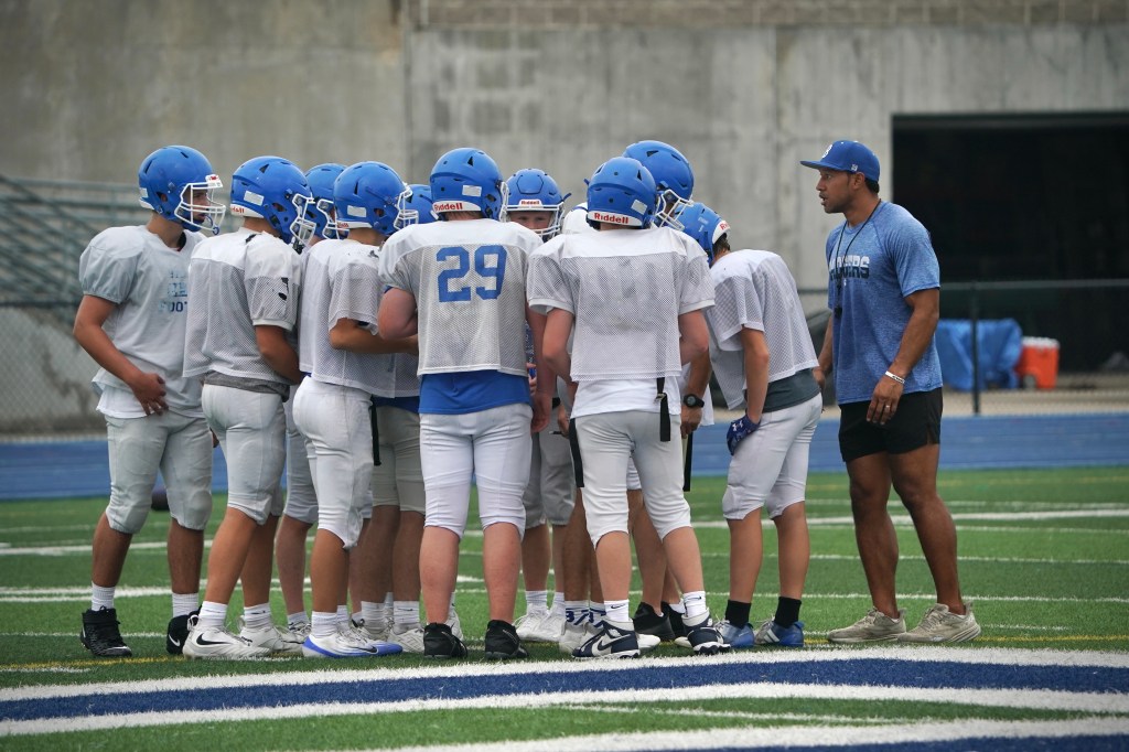 A group of young football players in blue helmets and practice jerseys huddled together on a field, with a coach providing guidance.