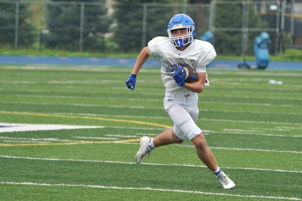 A young male football player in a white jersey and blue helmet running with a football on a green turf field during practice.