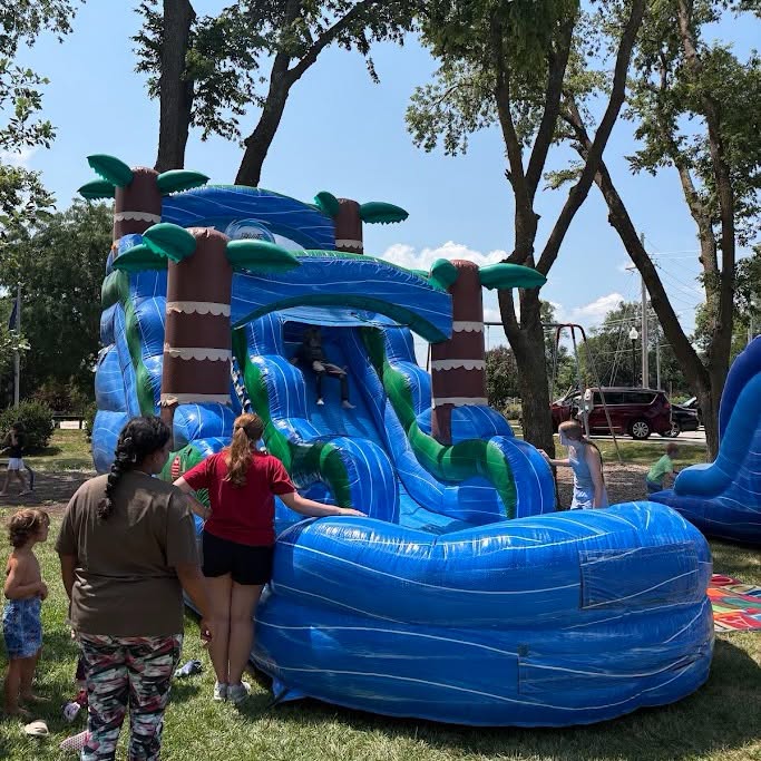 A vibrant inflatable water slide designed like a tropical scene, featuring palm trees and cascading blue slides, with children playing nearby in a sunny park.