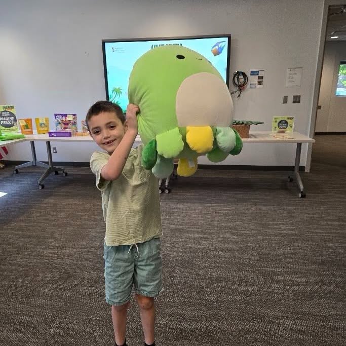 A young boy smiles while holding a large green plush toy in a room with a presentation screen and tables in the background.