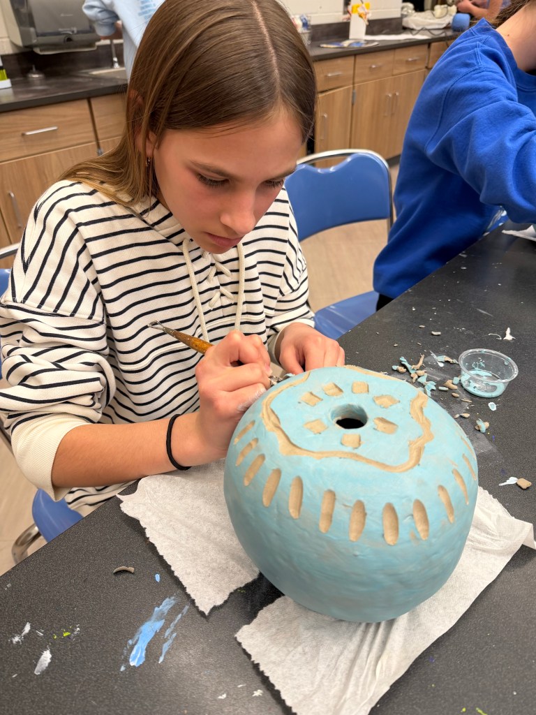 A young girl painting a blue pottery bowl with a brush, focused on her work. She is seated at a table with art supplies and tools around her.